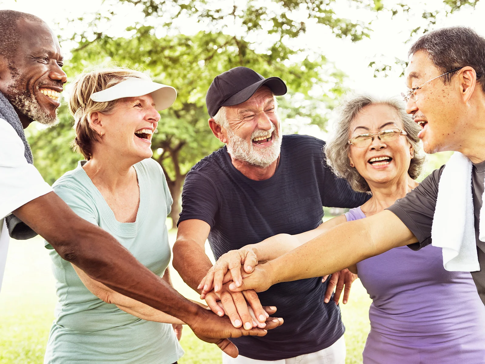 Diverse group of smiling adults stacking hands together outdoors in a park setting.